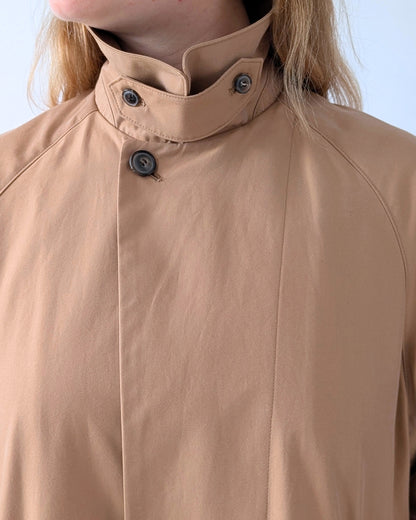 Close-up of a person wearing a beige high-neck jacket with a buttoned collar and dark buttons, against a light blue background.
