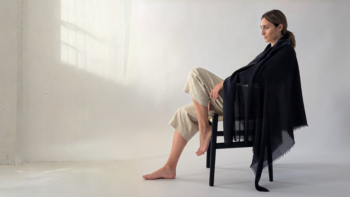 A woman sits barefoot on a black chair in a bright, minimalist studio, draped in a large black shawl or wrap over beige cropped pants.